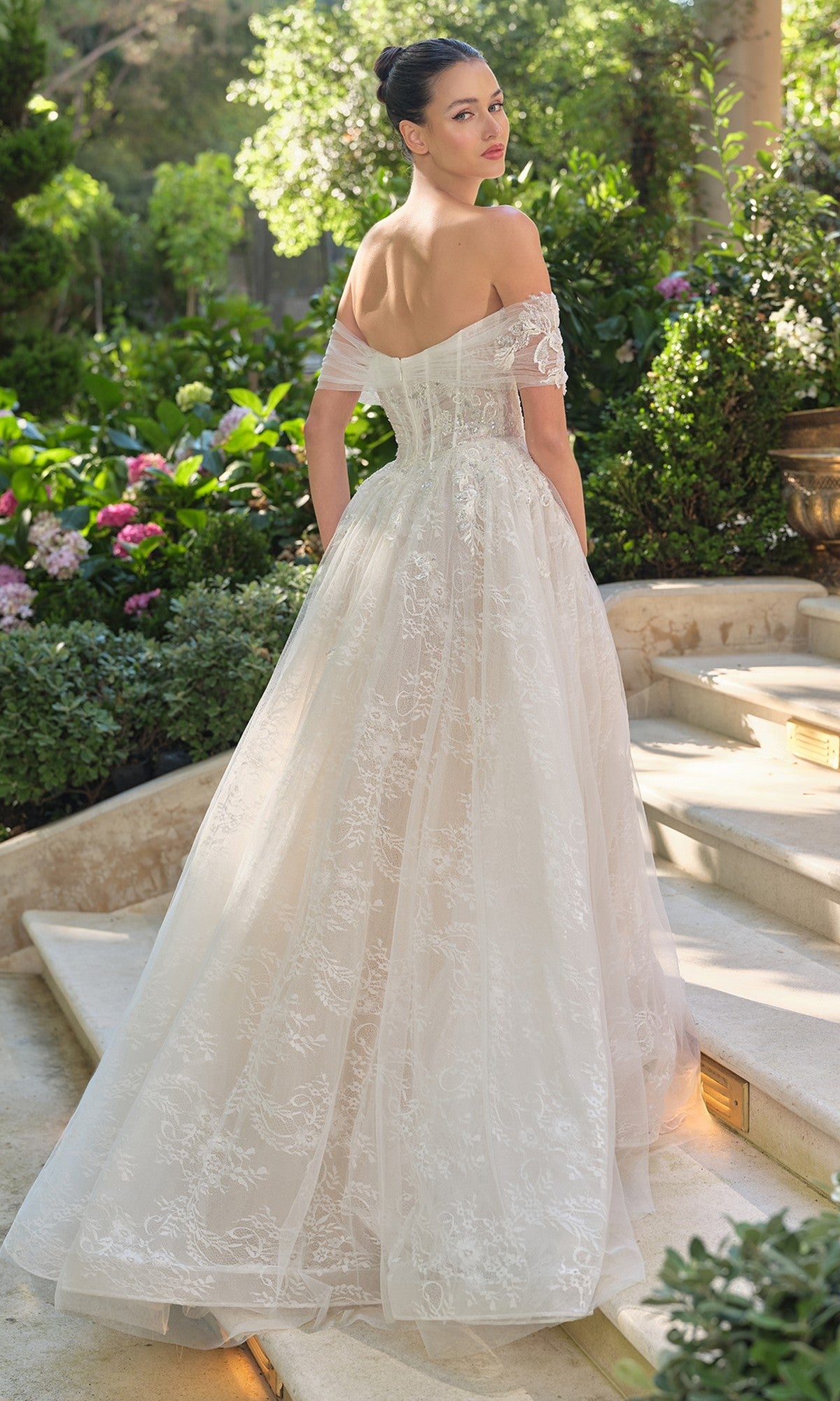 A woman wearing the Andrea and Leo WL090 long off-the-shoulder lace wedding dress with a corset bodice stands on stone steps in a garden, glancing back over her shoulder.